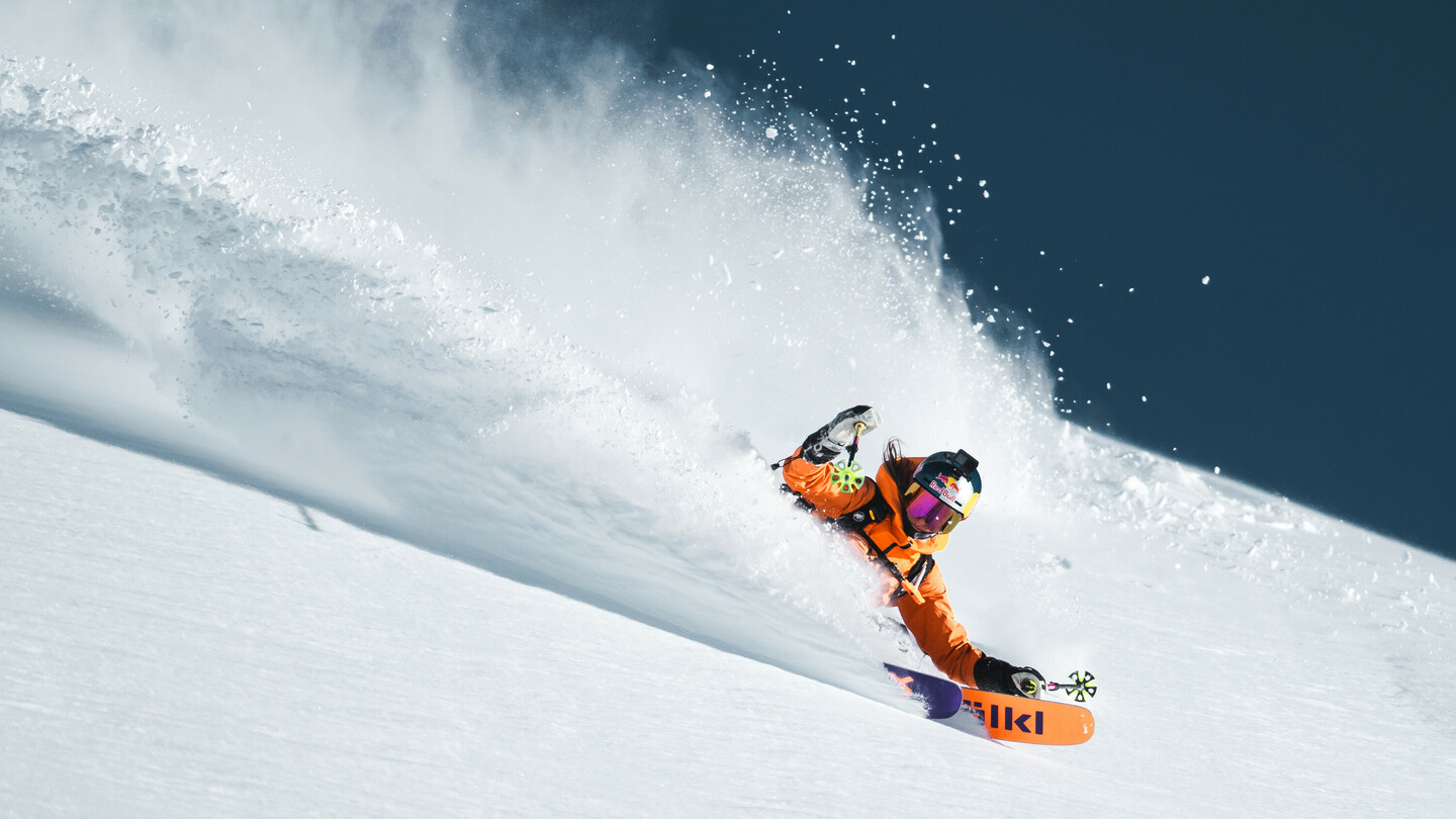 Female skier in an orange jacket carving dynamically through deep powder snow, with snow spraying in the background.