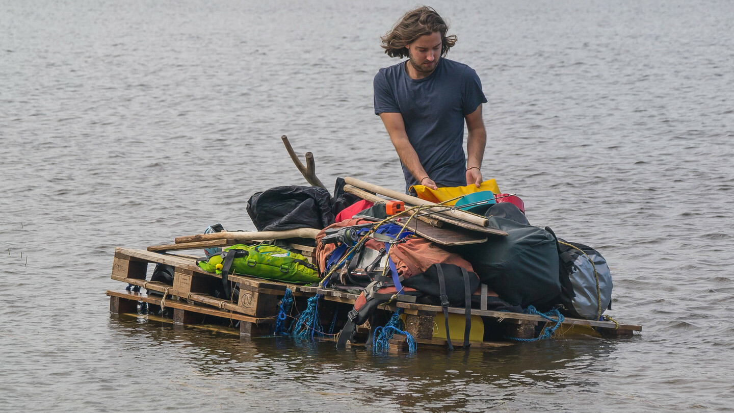 A man with shoulder-length hair stands on a makeshift raft made of wooden pallets on a lake. The raft is loaded with various items, including bags, paddles, and gear. In the background, a shore with dense forest is visible.